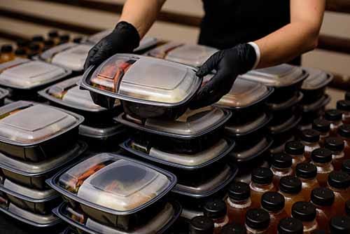 Catering worker wearing rubber gloves stacking plastic sectioned carry-out boxes filled with food for a catered event.