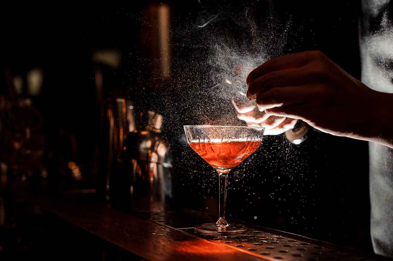 Bartender's hands sprinkling the juice into the cocktail glass filled with alcoholic drink on the dark background