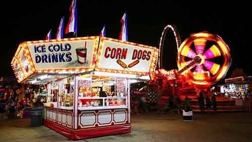 A concession stand at a carnival with lit signs for "ice cold drink" and "corn dogs." A carnival rid lit with yellow, red, and pink lights spins in the background.