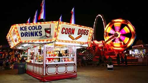 A concession stand at a carnival with lit signs for "ice cold drink" and "corn dogs." A carnival rid lit with yellow, red, and pink lights spins in the background.