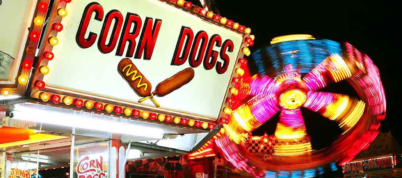 Illuminated corn dog sign on a concession stand and a fair with a carnival ride lit with red, yellow, pink, and blue lights spins in the background.