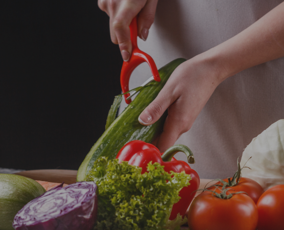 Cook using a y-shaped peeler to peel a cucumber. Other vegetables: red cabage, lettuce, red bell pepper and tomatoes in the foreground.