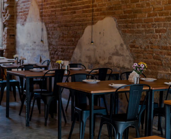 Photograph of a restaurant dining room with an unfinished brick wall, flat black stamped metal chairs and natural wood tables with flat black metal legs.