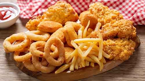 Deep fried onions, french fries, chicken nuggets, and a fried chicken on a wooden plate on a wood grain table. A ramakin of ketchup and a red and white checked table cloth in the background.