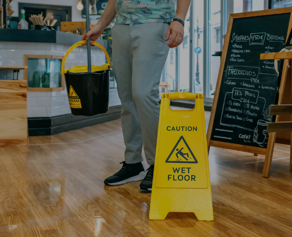 Person walking on wood floor with a mop bucket and mop in one hand as they walk by a yellow sign that says "Caution Wet Floor."