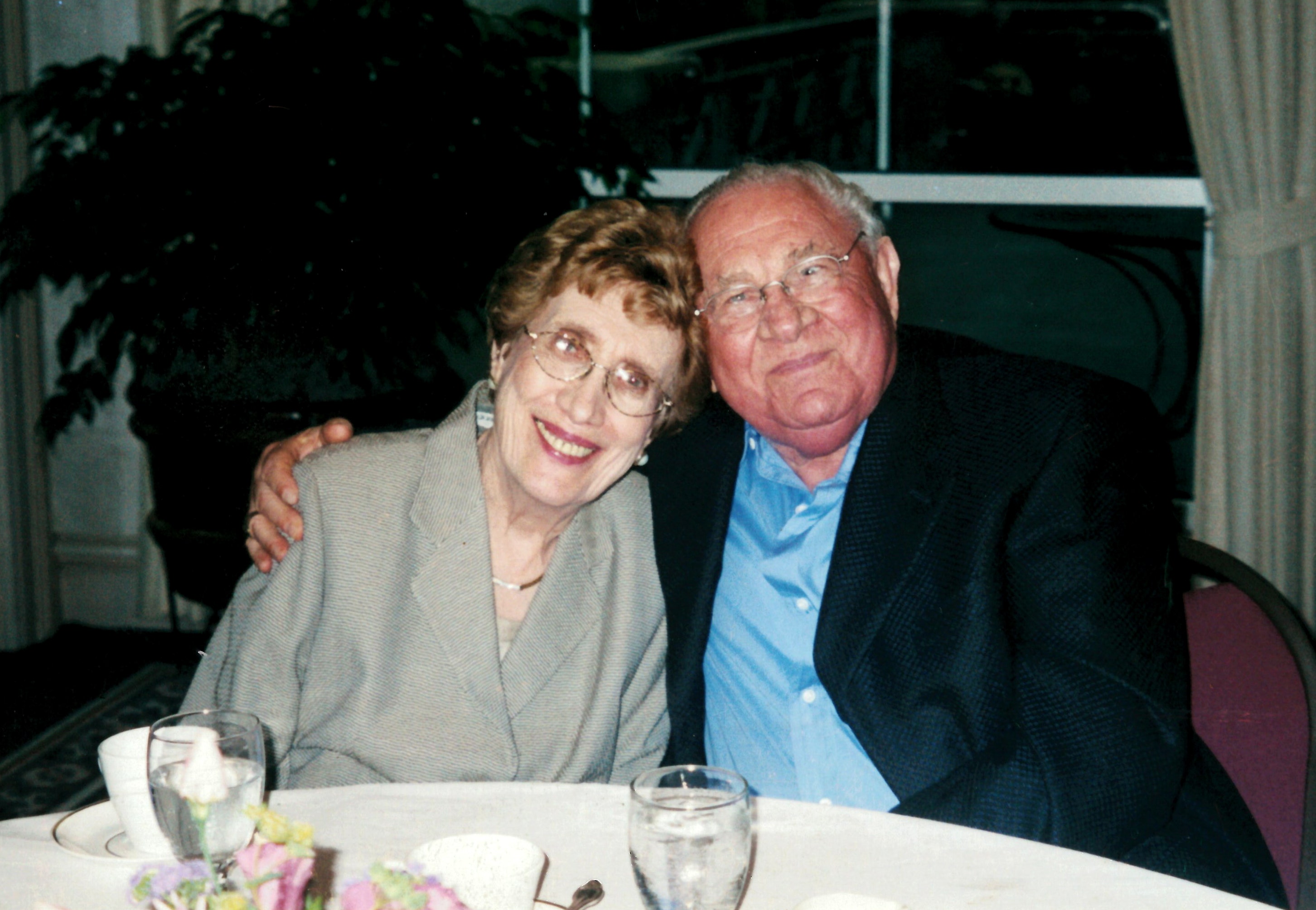 Elderly couple seated at a dinning table in a restaurant. Morris Dach, right and his wife Marian. Morris is wearing a blue dress shirt and black blazer. Marion is wearing a light grey jacket.