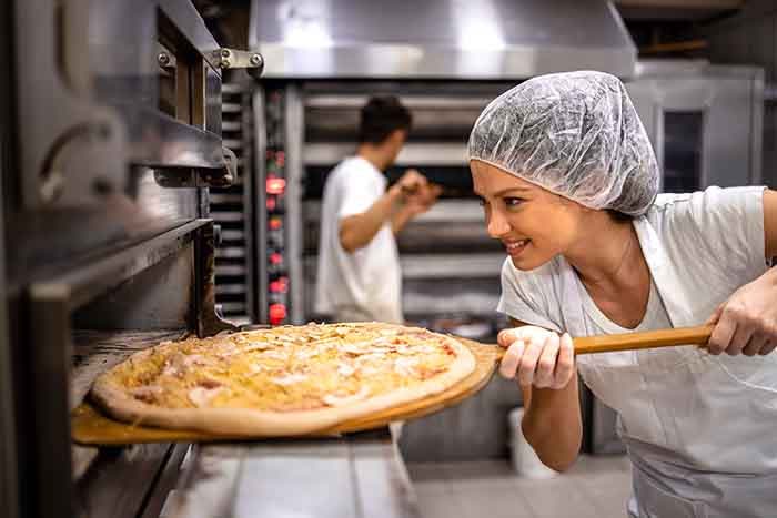 Female chef in white uniform and hairnet putting pizza in the oven for baking process.