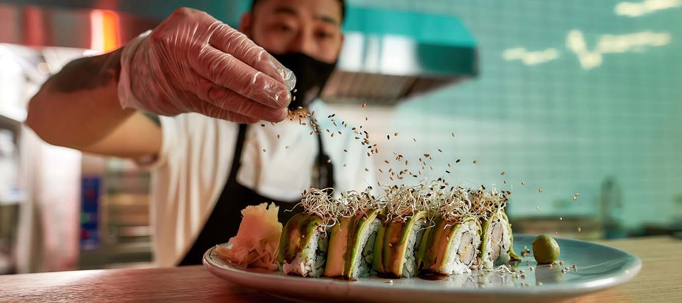 Masked chef standing in the kitchen and sprinkles sesame seeds on sushi on the table.