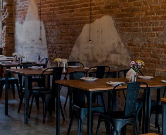Photograph of a restaurant dining room with an unfinished brick wall, flat black stamped metal chairs and natural wood tables with flat black metal legs.