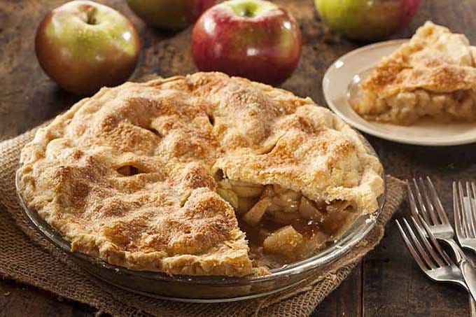 Double crust apple pie with a piece missing in a glass pie dish sitting on a square of burlap. Red apples in background and a piece of pie sitting on a white plate. The scene is set on a wooden table.