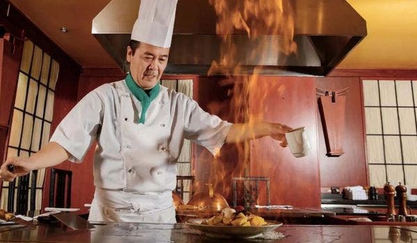 Asian chef performing a dramatic flaming teppanyaki cooking technique in a restaurant kitchen.