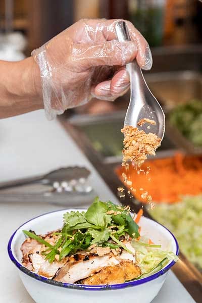 Chef preparing fresh grilled chicken noodle bowl on the cutting board of a sandwich prep table.