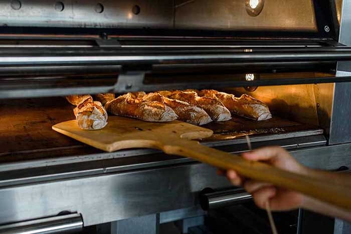 Close up of baker is taking off from oven the french baguette bread with wood peel at commercial bakery.