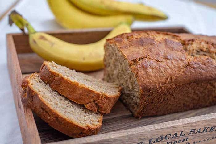 A loaf of banana bread sits in a wooden crate with bananas in the background.