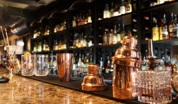 cocktail glasses and shakers on a marble bartop with liquor bottles on shelves in the background.