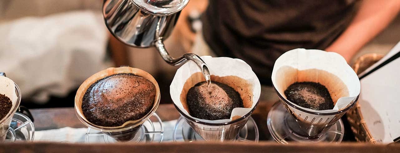 A barista uses a stainless steel hot water pot to pour water over coffee grounds in filter to make pour over coffee.
