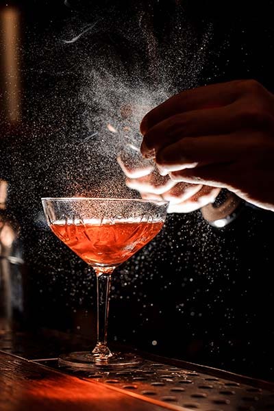 Bartender's hands sprinkling the juice into the cocktail glass filled with alcoholic drink on the dark background