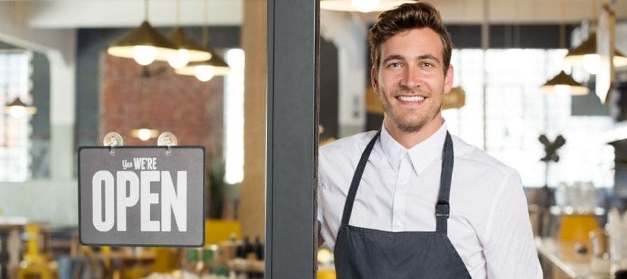 White man in white button shirt with collar wearing a navy blue apron standing in the doorway of a cafe with a "Yes, we're open" sign in the window next to him.