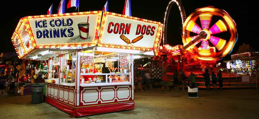 Food stand lit up with colored lights at a carnival at night. Lit up amusement rides in background with yellow and red lights.