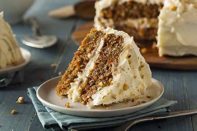 A slice of carrot cake on a small white plate sitting on folded blue cloth napkin with the cut cake in the background.