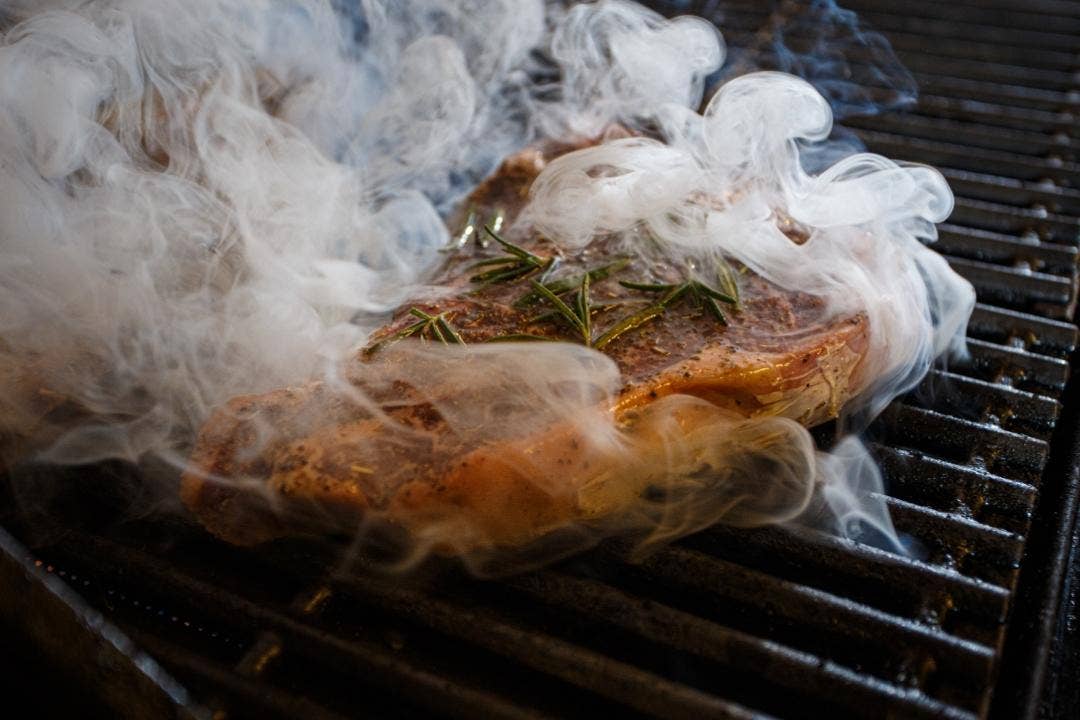 Juicy steak smoking on charbroiler grill with rosemary sprigs.