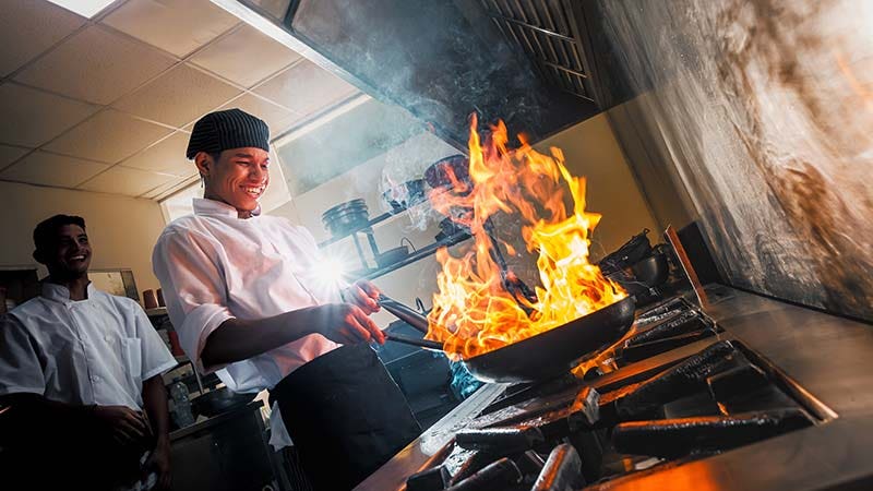 Young chef in the restaurant kitchen cooking with flames in a fry pan.