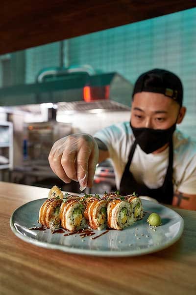 Professional sushi chef wearing protective mask and gloves decorating sushi rolls preparing to be served on plate at commercial kitchen.