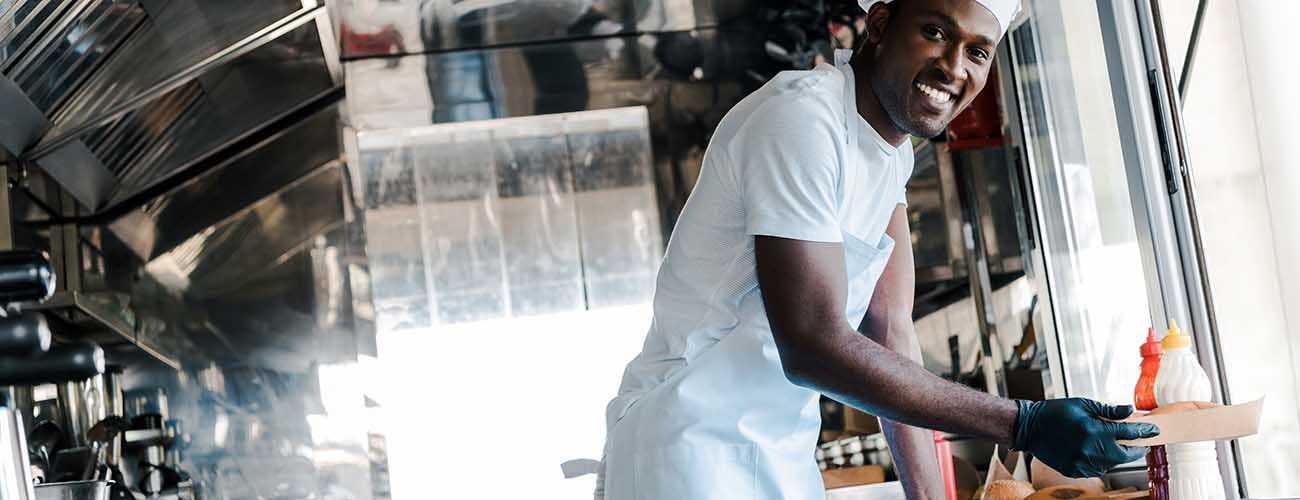selective focus of cheerful black chef smiling while holding carton plate in food truck
