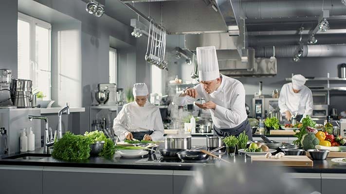 Chef works in a big restaurant kitchen with his help. Kitchen is full of food, vegetables and boiling dishes. He is taste food.