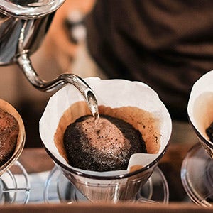 A barista uses a stainless steel hot water pot to pour water over coffee grounds in filter to make pour over coffee.