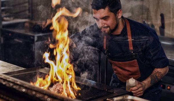 Chef grilling meat on a charbroiler with visible flames in a commercial kitchen.