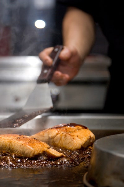 Two pieces of salmon on a griddle with a cook waiting with a offset spatula.