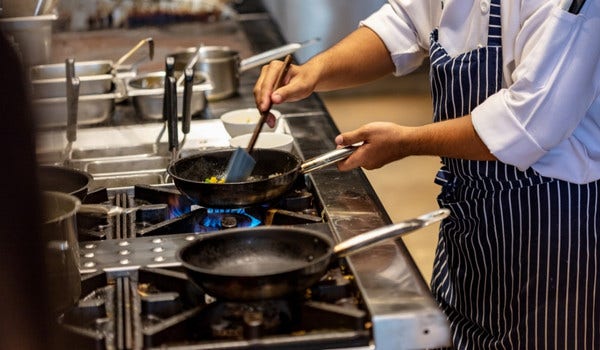 Chef cooking on a commercial gas stove in a professional kitchen with frying pans and utensils.