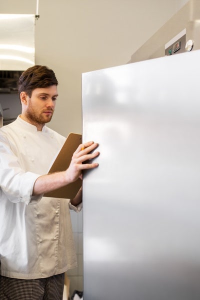 Male chef looking into a reach-in commercial refrigerator holding a clipboard.