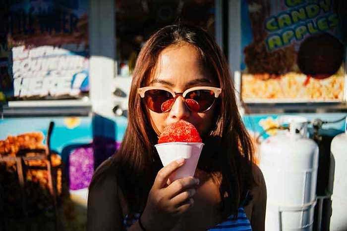 A young woman with sunglasses on eats a snow cone in front of a concession stand.