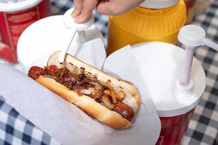 Ketchup being pumped from a condiment dispenser onto a grilled onion covered hot dog, with a checkered tablecloth in the background.