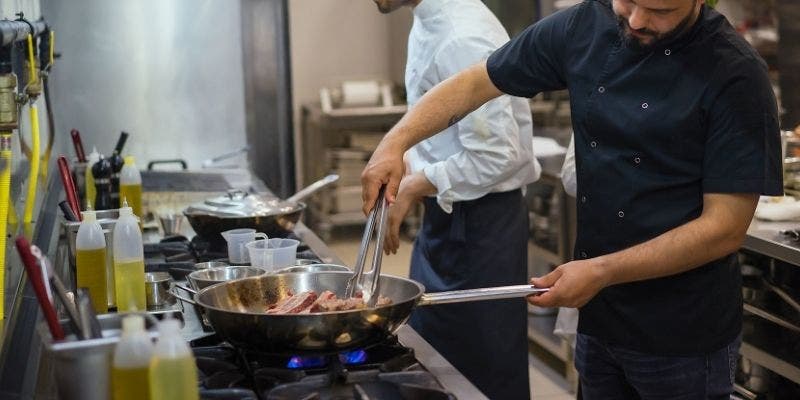 Two male chefs, one in a white a chefs jacket and the other in a black. Cooking beef in a stainless steel skillet.