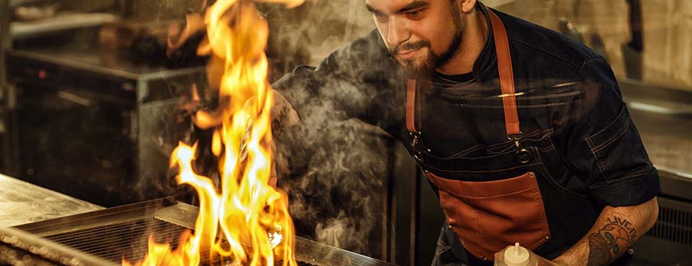 Bearded chef in denim apron and with tattoo on forearm holding oil in hand cooking meat on grill with fire and smoke.