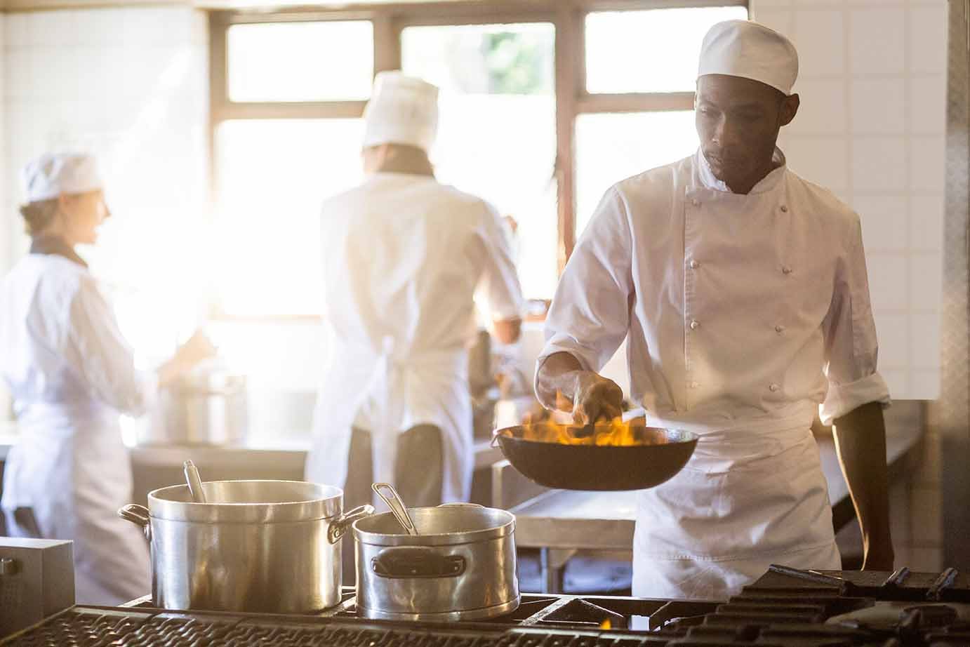 Professional cook holding fry pan with flaming contents in backlight commercial kitchen. Two cooks are talking to each other in the background.