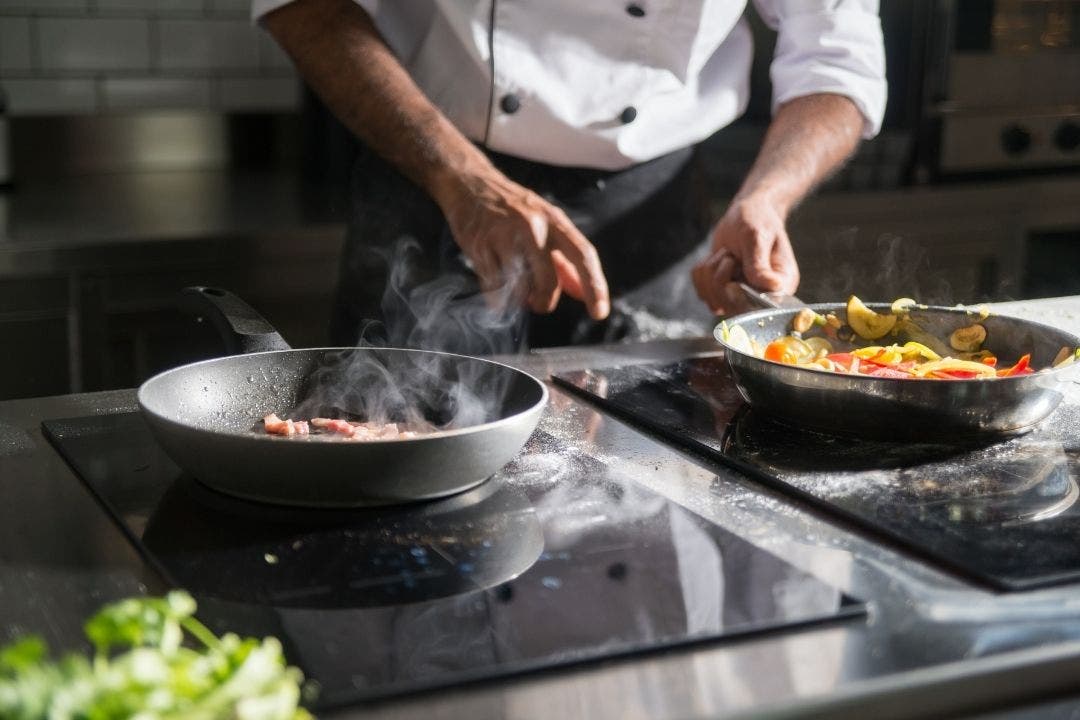 Chef sautéing vegetables and meat in skillets on induction cooktop