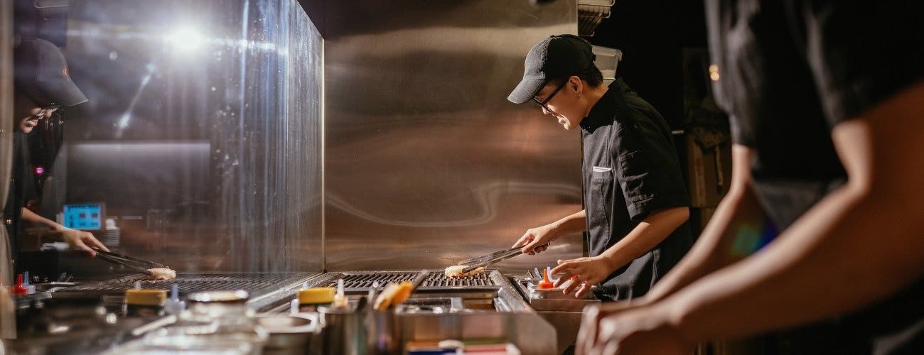 Two Asian male chefs in black chefs coats preparing food on a comemrcial kitchen line usining a charbroile.
