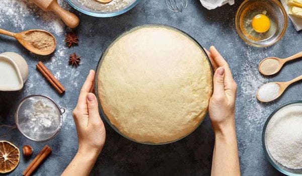 Hands holding a bowl of freshly risen dough surrounded by baking ingredients on a countertop.