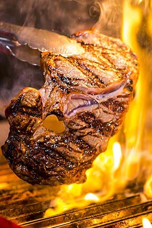 Close up of chef using tongs to flip steak on a charbroiler as flames shoot up around it.