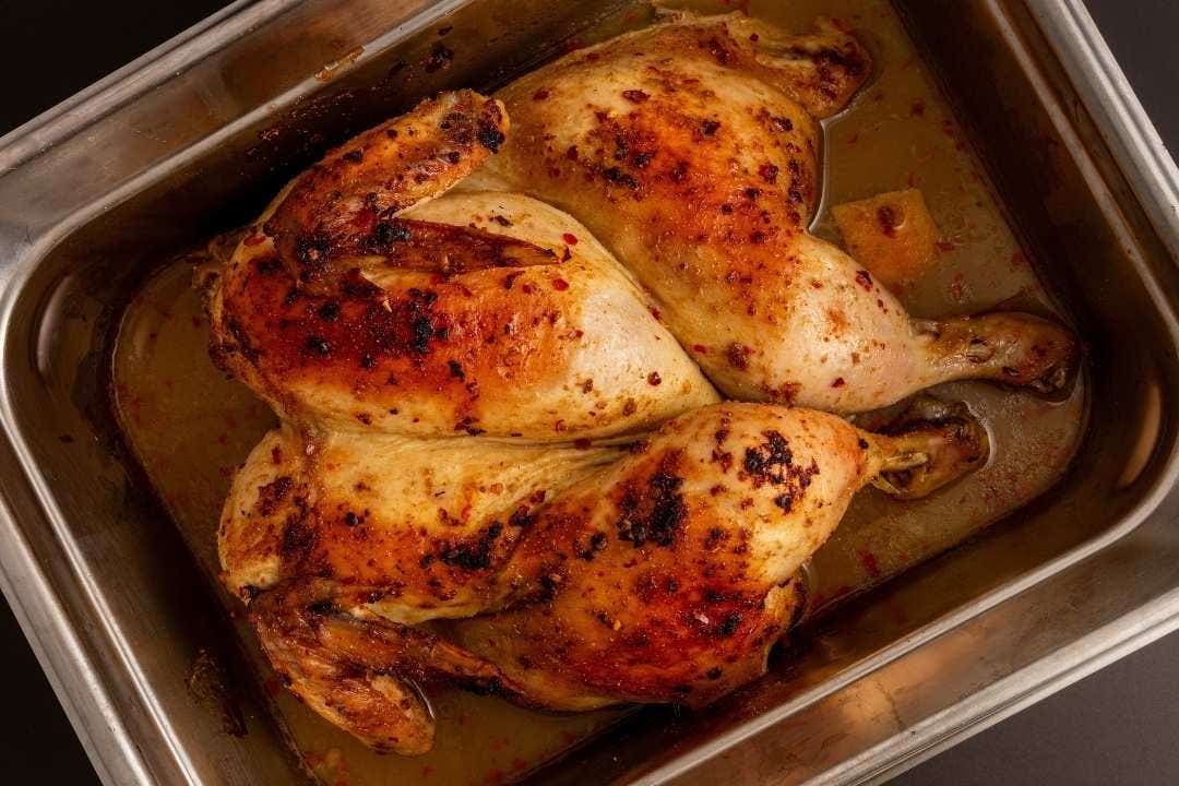 Whole roasted chicken in a stainless steel food pan with juices and seasoning