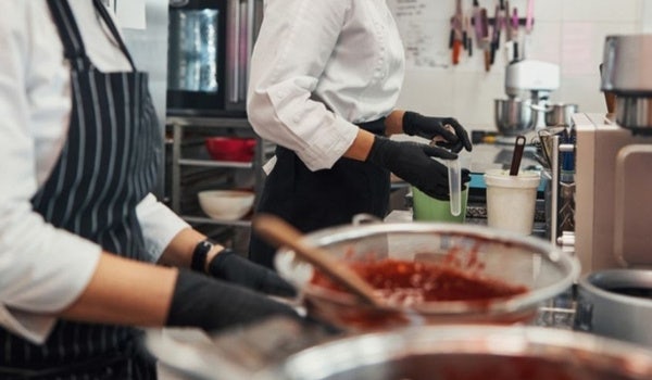 Chefs in a professional kitchen preparing food with mixing bowls and utensils, wearing black gloves and aprons.