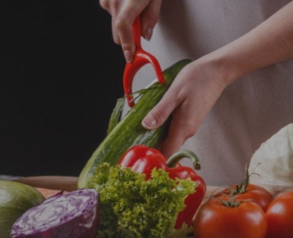 Cook using a y-shaped peeler to peel a cucumber. Other vegetables: red cabage, lettuce, red bell pepper and tomatoes in the foreground.