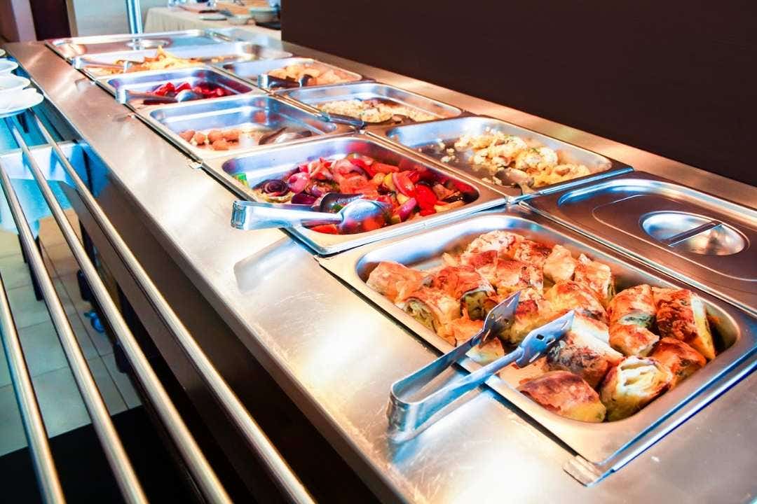 Hot food items displayed in stainless steel pans at a school cafeteria buffet