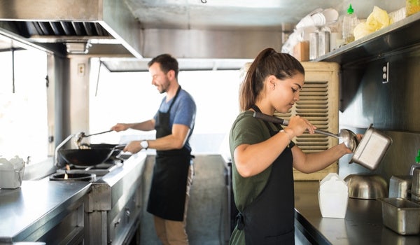 Male and female cooks inside of a food truck making food during a sunny day.