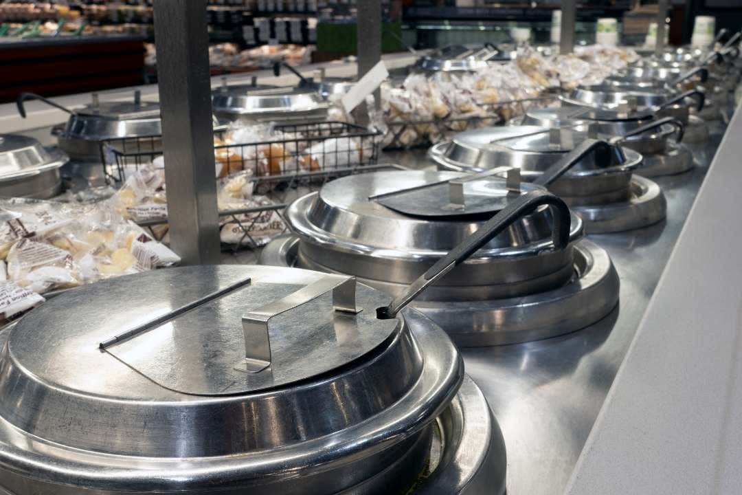Row of stainless steel soup warmers with lids and ladles in a grocery hot bar section