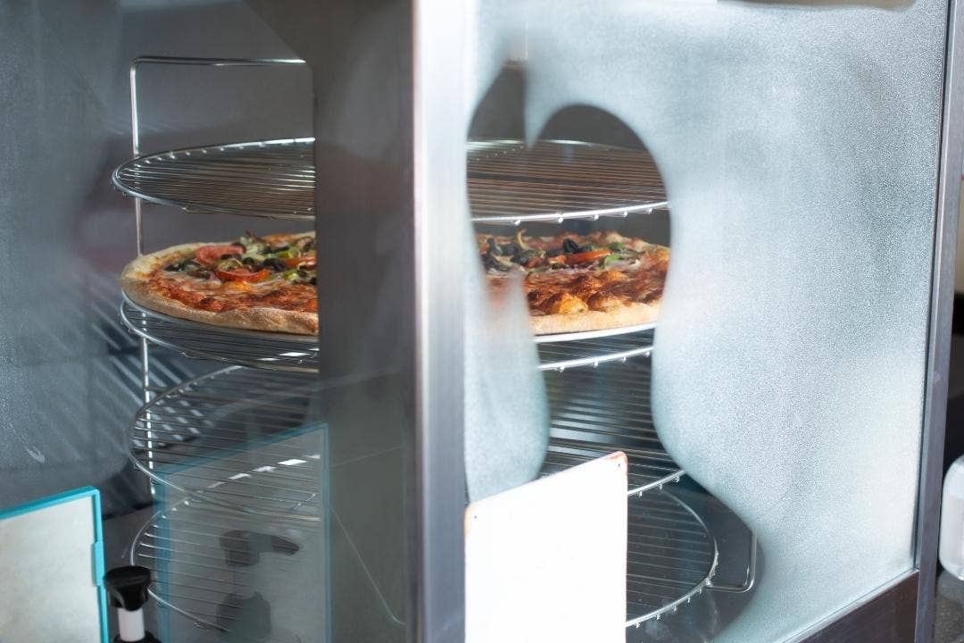 Fresh pizzas displayed inside a heated rotating pizza warmer with glass doors